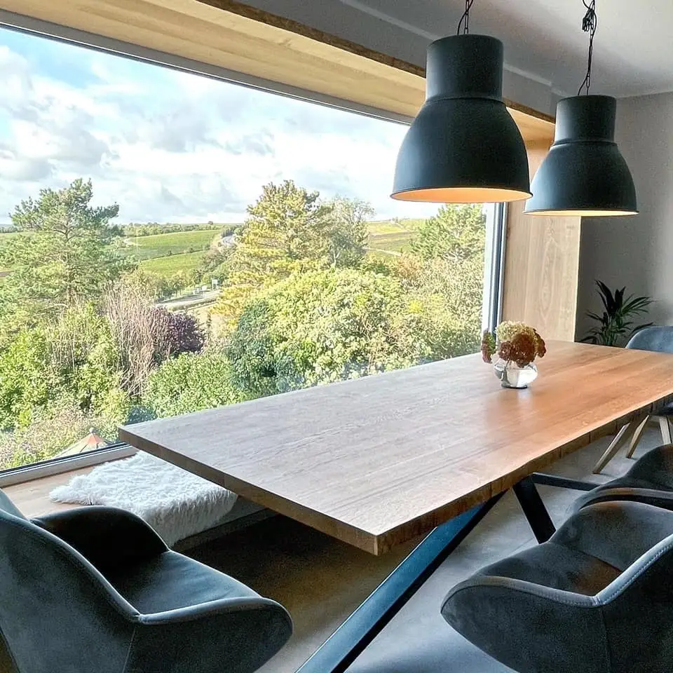 Essbereich mit großem Holztisch aus massiver Eiche mit Baumkante, modernen schwarzen Hängelampen und großem Sitzfenster mit Ausblick auf die Natur.