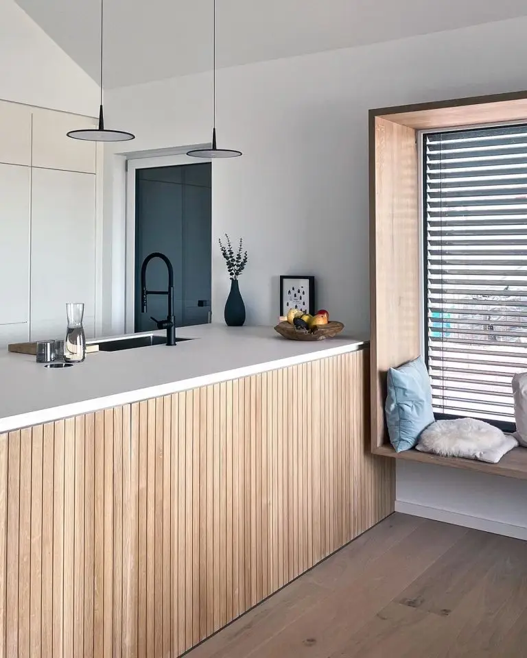 Moderne Küche in GERBERHAUS7 mit heller Kochinsel, integriertem schwarzen Wasserhahn, zwei Hängeleuchten und einladendem Sitzfenster mit Kissen.