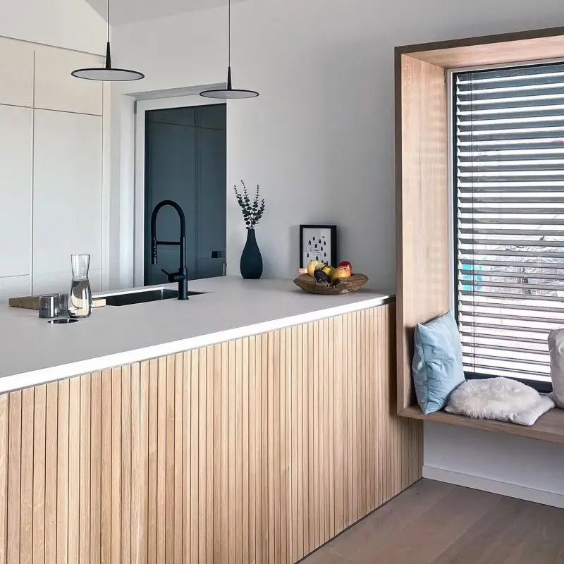 Moderne Küche in GERBERHAUS7 mit heller Kochinsel, integriertem schwarzen Wasserhahn, zwei Hängeleuchten und einladendem Sitzfenster mit Kissen.