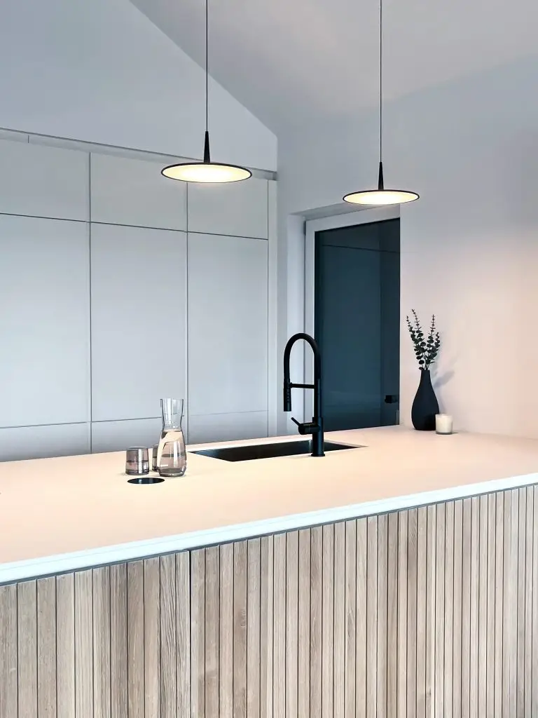 Küche mit weißer Kochinsel, Holzfronten, schwarzem Wasserhahn und zwei modernen Hängeleuchten.