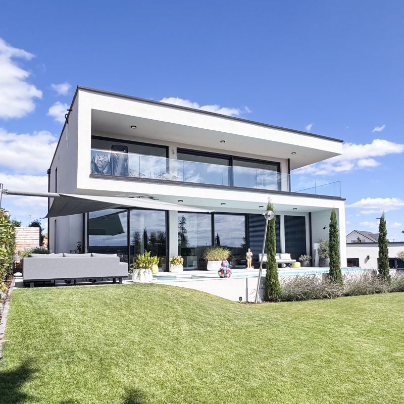 Moderne Stadtvilla mit Walmdach, großer Glasfassade, Poolterrasse und weitläufigem Garten unter blauem Himmel