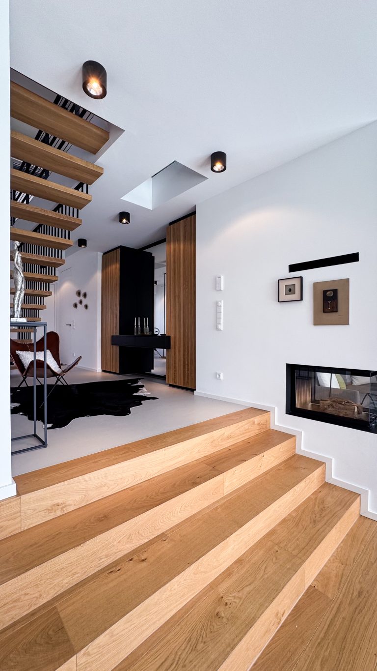 Innenraum im #GERBERHAUS12 mit Eichenholzstufen, freitragender Holztreppe mit schwarzen Stahlstreben, eingebautem Kamin und schwarzen sowie holzverkleideten Einbaumöbeln.