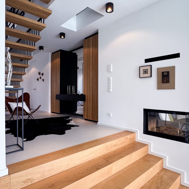 Innenraum im #GERBERHAUS12 mit Eichenholzstufen, freitragender Holztreppe mit schwarzen Stahlstreben, eingebautem Kamin und schwarzen sowie holzverkleideten Einbaumöbeln.