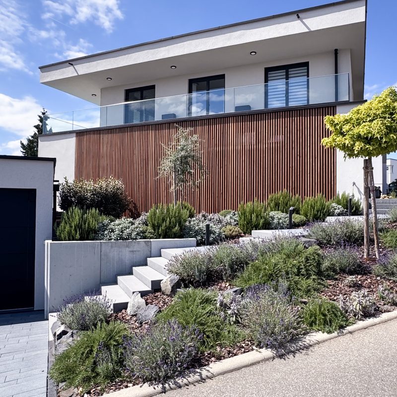 Außenansicht des #GERBERHAUS12 mit großer Glasbalustrade, weißer Putzfassade, vertikaler Holzlamellenverkleidung und terrassierter Hanggestaltung aus Naturstein, Lavendel und mediterranen Pflanzen.