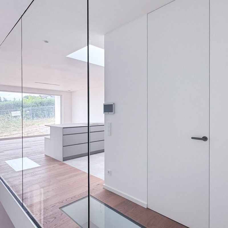 Innenansicht im #GERBERHAUS16 mit bodentiefer Glaswand, begehbarem Glaselement im Eichenboden und Blick in den offenen Küchenbereich mit großem Fenster.