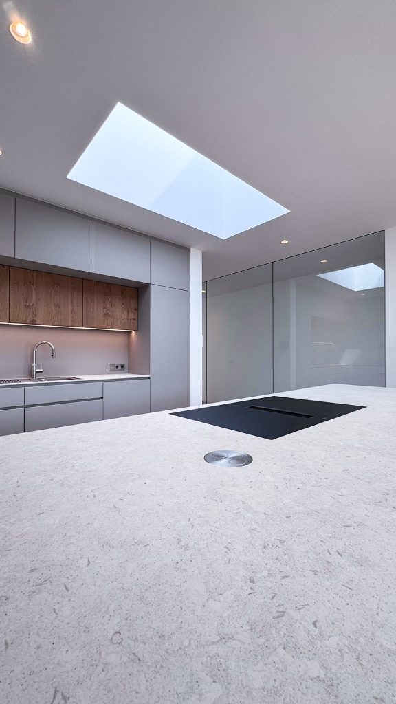 Innenansicht im #GERBERHAUS16 mit moderner Kücheninsel, grauen Fronten, Holz- und Steindetails sowie großem Oberlicht für natürliche Beleuchtung.