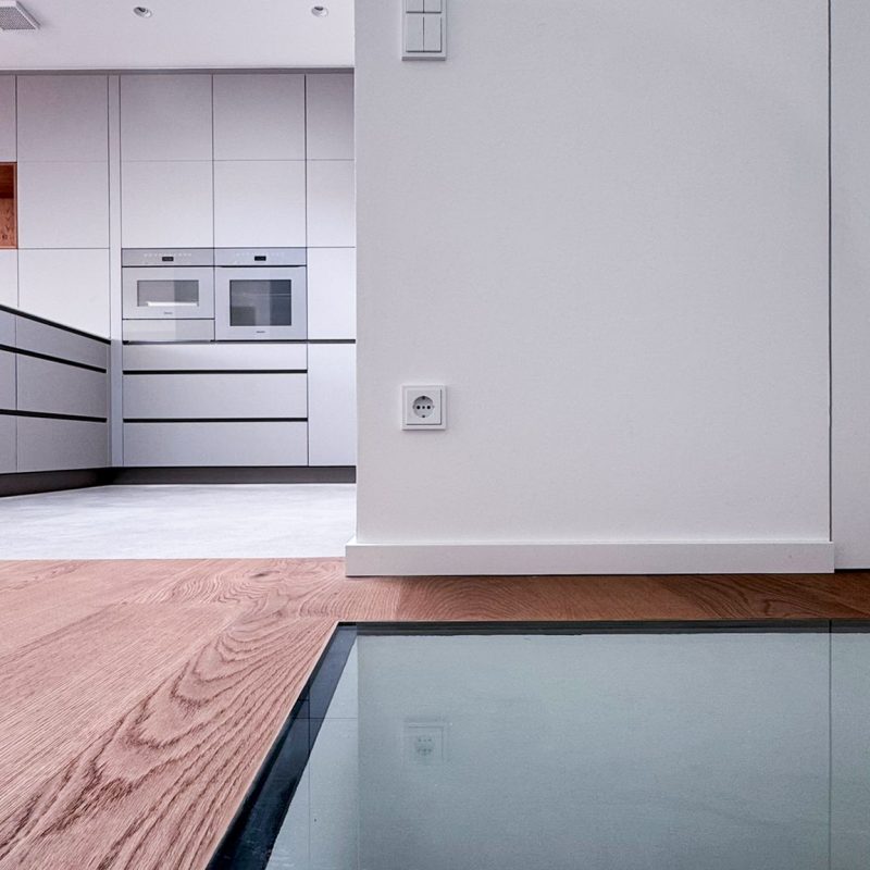 Glasboden im Wohnbereich mit Blick in darunterliegenden Raum, Parkettboden und Sicht auf moderne Küche mit Miele Geräten in Pearl Beige in #GERBERHAUS16