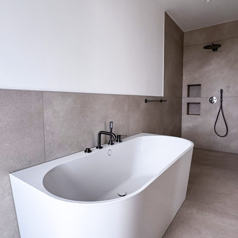 Modernes Badezimmer im #GERBERHAUS16 mit freistehender weißer Badewanne, beiger Großformatfliese, schwarzer Armatur und bodengleicher Walk-In-Dusche mit integrierten Nischen.