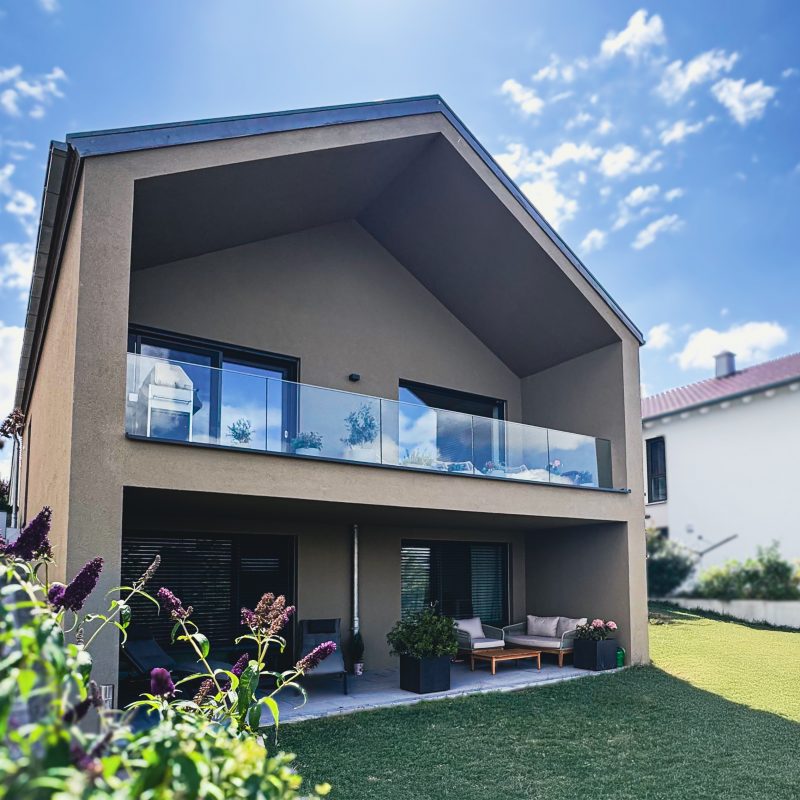 Außenaufnahme von #GERBERHAUS7 mit überdachtem Balkon mit Glasgeländer, offener Terrasse im Erdgeschoss und gepflegtem Garten.