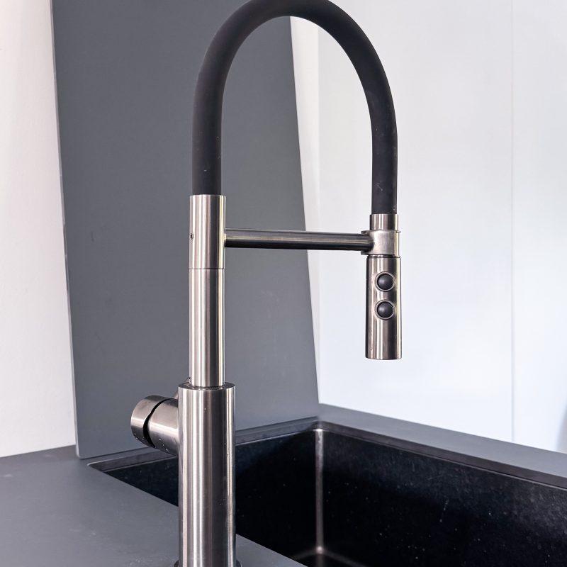 Detailaufnahme einer modernen Edelstahl-Küchenarmatur mit schwarzem Flex-Schlauch und Bedienknöpfen, eingebaut in eine dunkle Spüle im Umbau U06.
