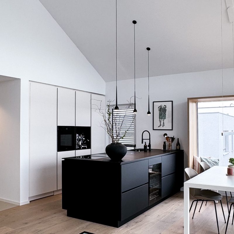 Offene Küche mit schwarzer Kücheninsel, modernen Hängeleuchten und verstecktem Stauraum, kombiniert mit weißen Wänden und einem Essbereich.