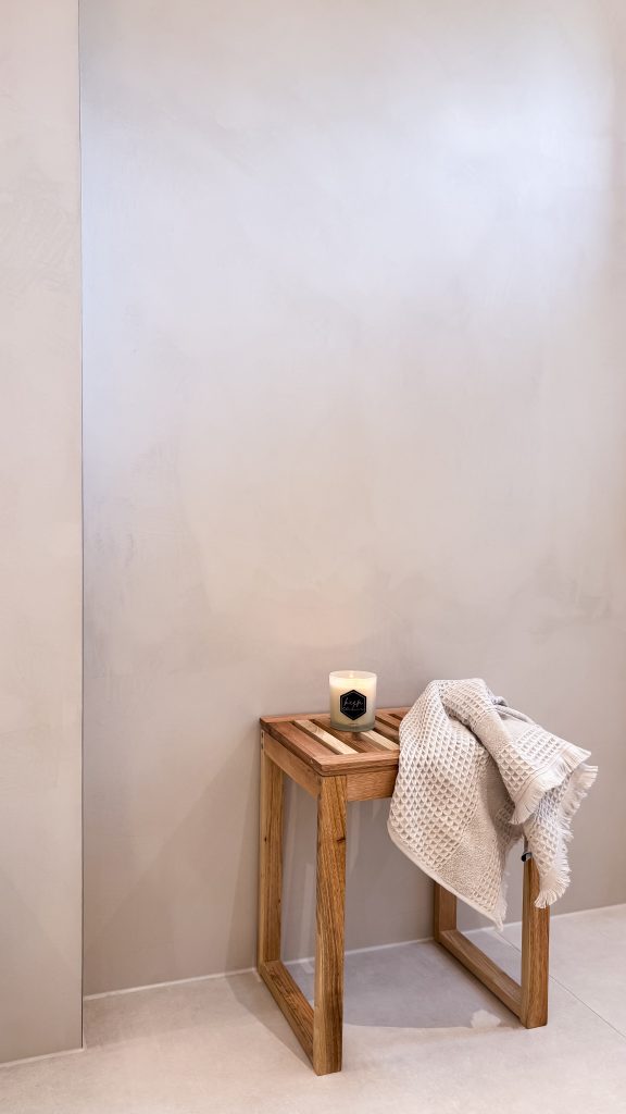 Minimalistisches Bad-Detail mit kleinem Holzschemel, Duftkerze und gefalteten Handtüchern vor einer mit Microzement verputzten Wand.