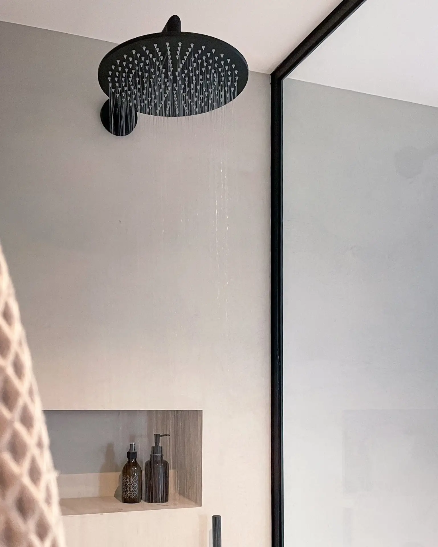 Schwarzer großer Regendusche Duschkopf an einer glattverputzten Wand in Betonoptik. Wasser läuft. Kleine Nische mit zwei braunen Glaspumpspendern. Schwarz eingefasste Duschwandabtrennung aus Glas.