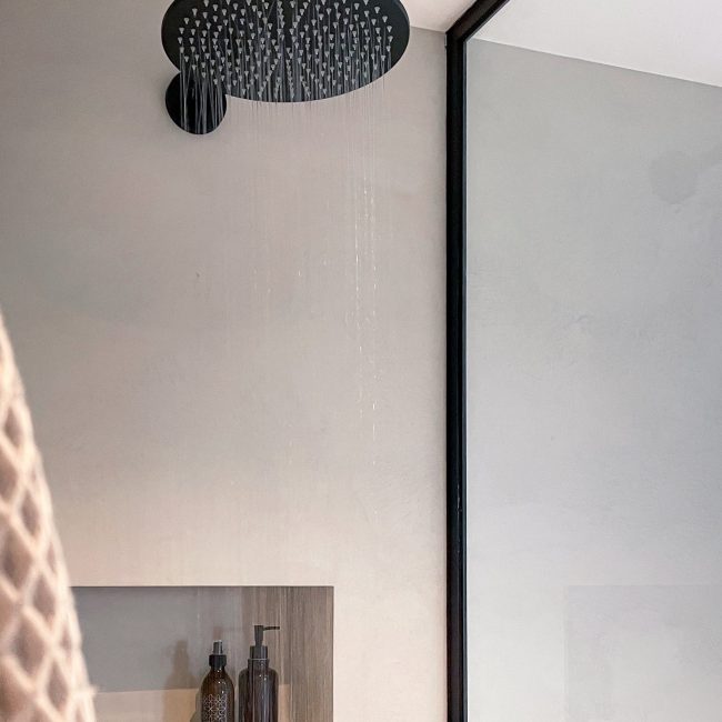 Schwarzer großer Regendusche Duschkopf an einer glattverputzten Wand in Betonoptik. Wasser läuft. Kleine Nische mit zwei braunen Glaspumpspendern. Schwarz eingefasste Duschwandabtrennung aus Glas.