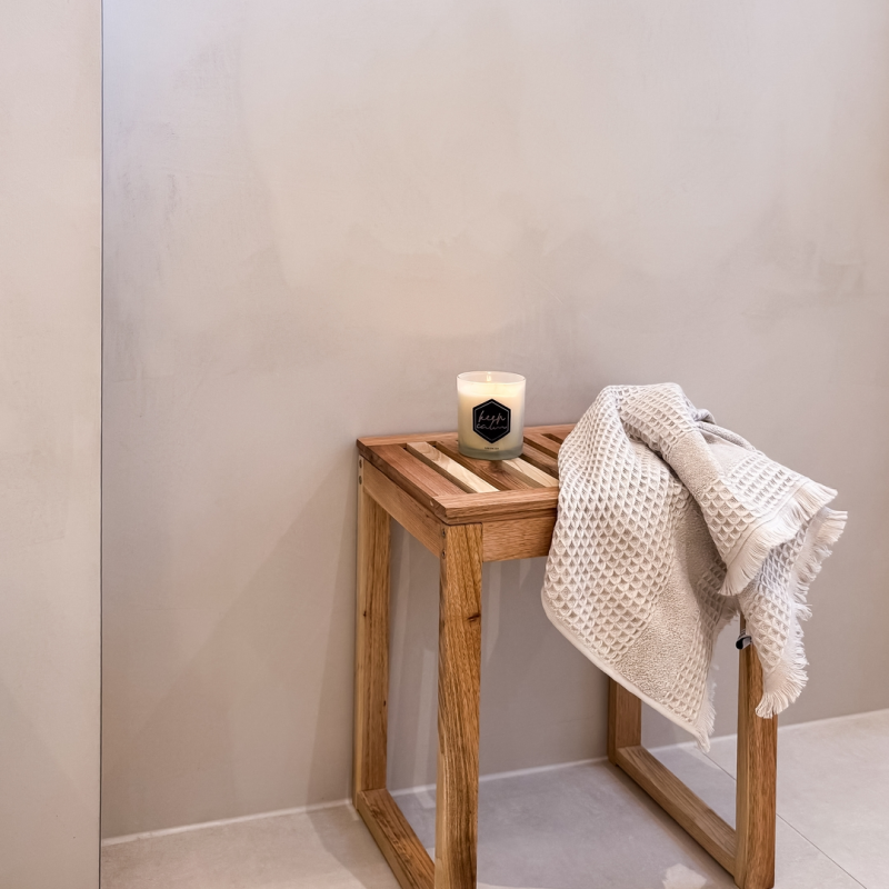 Minimalistisches Bad-Detail mit kleinem Holzschemel, Duftkerze und gefalteten Handtüchern vor einer mit Microzement verputzten Wand.