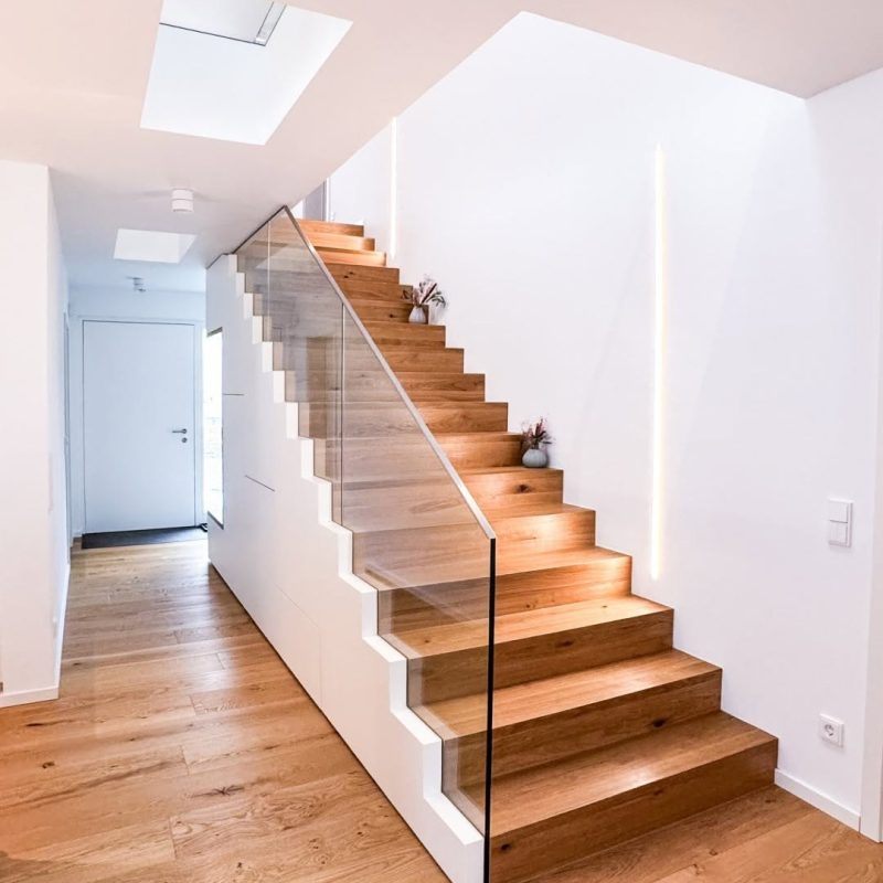 Treppe mit Stufen aus Eiche, rahmenlosem Glasgeländer, weißen Einbauschränken darunter und senkrechter LED-Lichtleiste im GERBERHAUS15