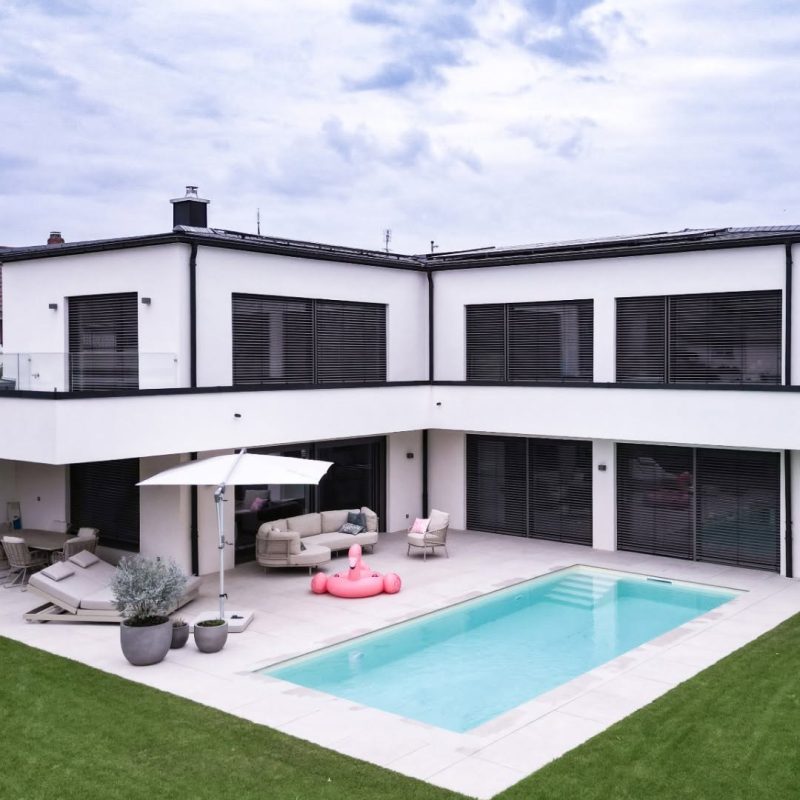 GERBERHAUS15 von oben: Einfamilienhaus in L-Form mit weißer Putzfassade, Terrasse mit Lounge, Pool und Rasen