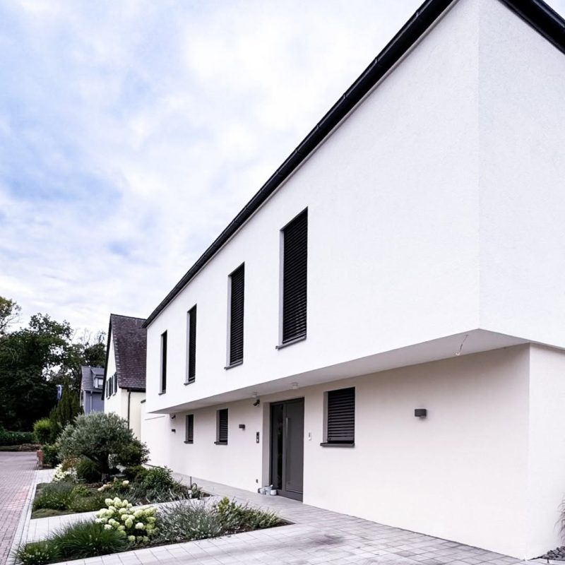 Straßenansicht des GERBERHAUS15: weiße Putzfassade mit auskragendem Obergeschoss, schmalen hohen Fenstern mit Raffstores, Eingangstür und Vorgarten mit Hortensien