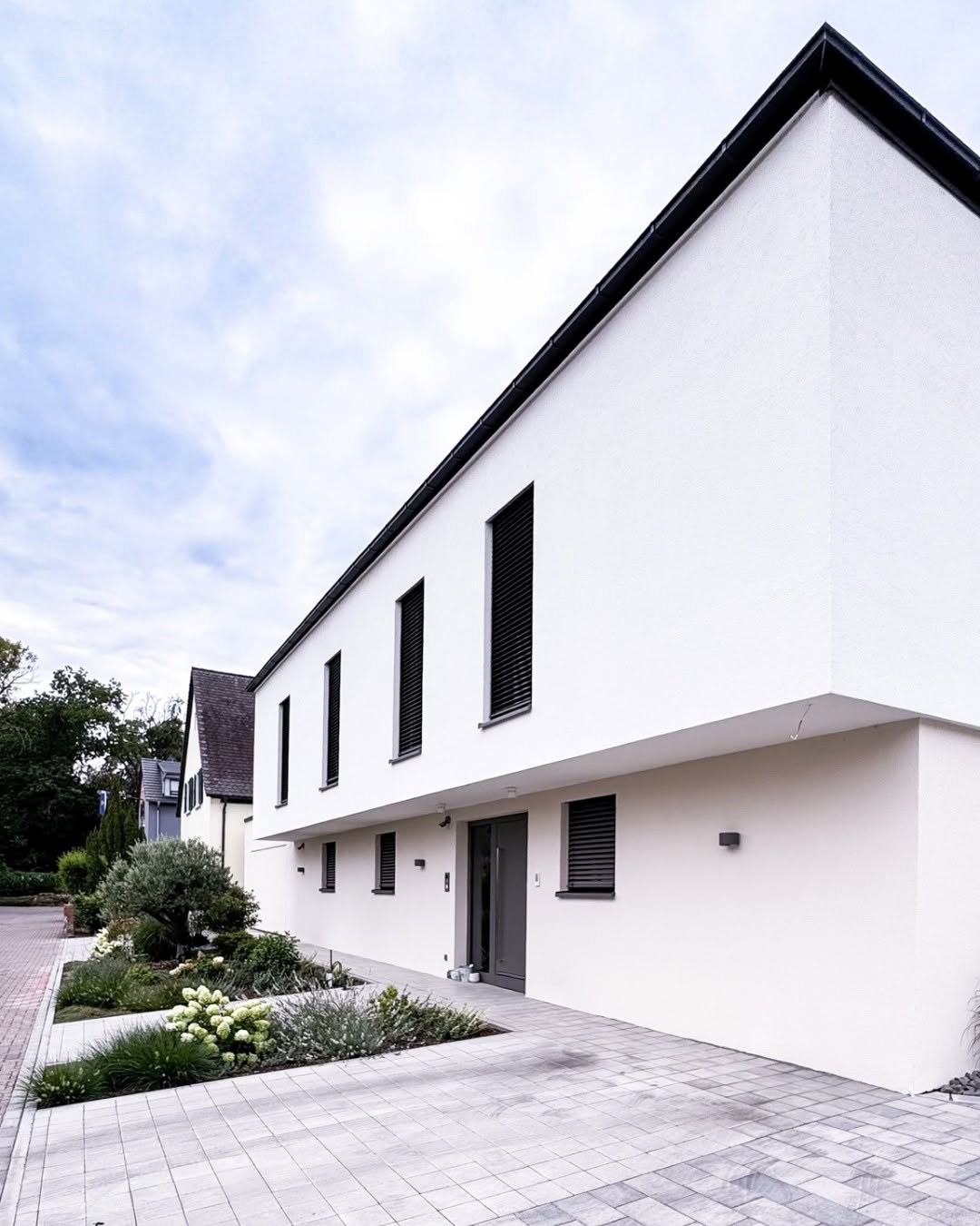 Straßenansicht des GERBERHAUS15: weiße Putzfassade mit auskragendem Obergeschoss, schmalen hohen Fenstern mit Raffstores, Eingangstür und Vorgarten mit Hortensien