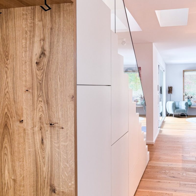 GERBERHAUS15: Sitznische mit Holzverkleidung, weißer grifflos eingebauter Treppenschrank und Glasgeländer im Flur