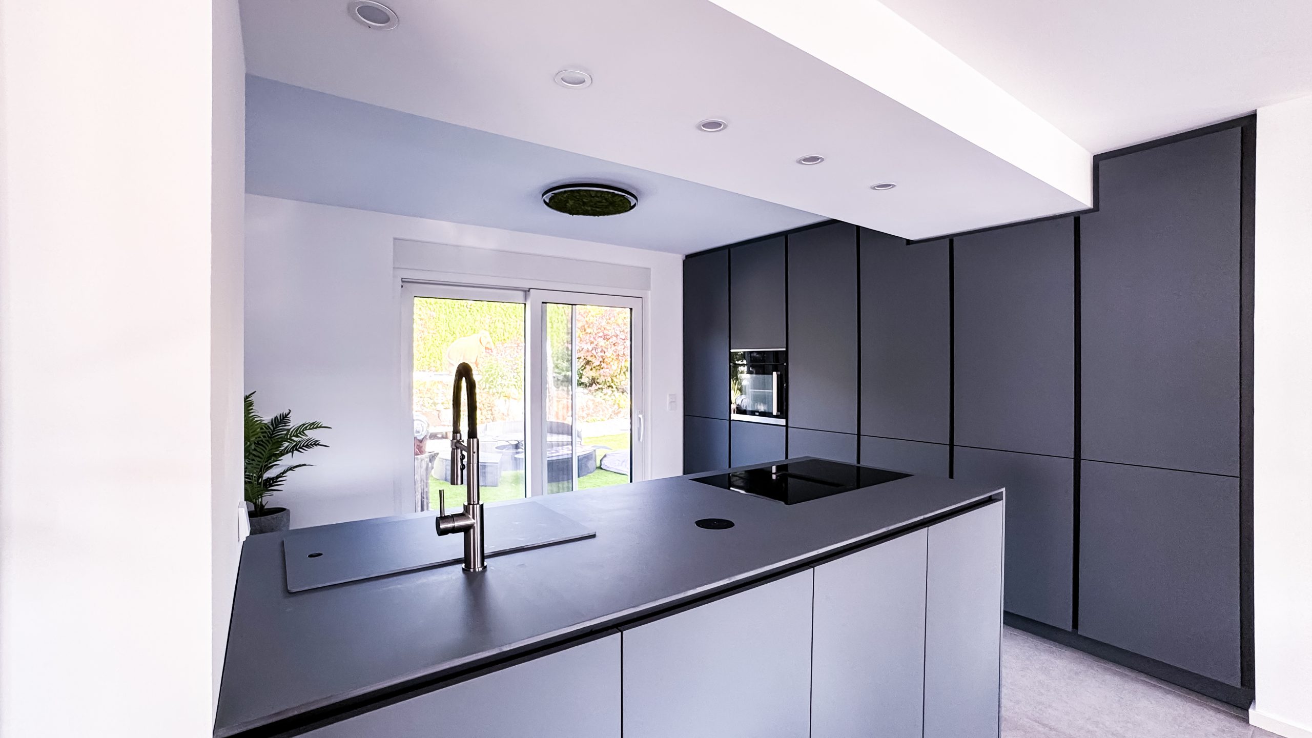 Innenansicht einer modernen Küche nach Umbau: schwarze Küchenfronten mit griffloser Optik, Kücheninsel mit Spüle und Induktionskochfeld, große Schiebetür zum Garten.